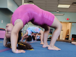 Kids in colorful outfits doing backbends on a blue gym mat in a gymnasium, with a focus on a girl in pink. Bright and lively scene.