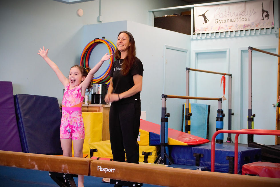 Girl in pink leotard on balance beam, arms raised in excitement, with smiling coach in gym. "Pathways Gymnastics" sign in background.