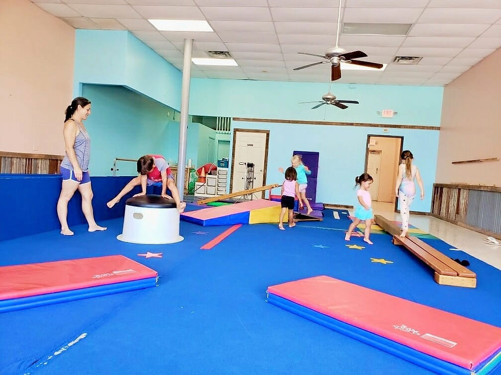Children play in a colorful indoor gym with mats, beams, and a drum. A woman watches. Bright blue floor, light walls, and fans overhead.