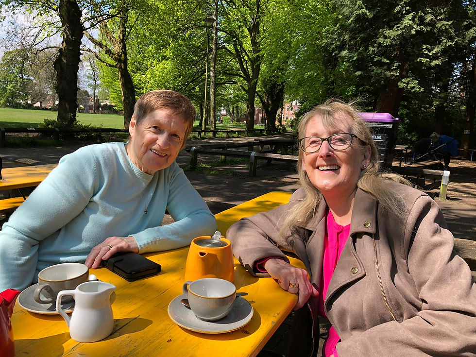 Two ladies in their early 60s sit at a picnic bench on a sunny day. They are smiling at the camera with a cup of tea each in front of them.