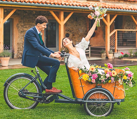 Le marié pédalant un vélo cargo fleuri avec la mariée, moment joyeux d’un mariage champêtre