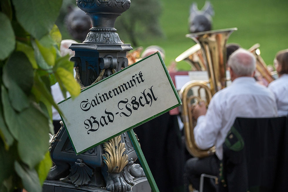 Ein Schild mit der Aufschrift „Salinenmusik Bad Ischl“ hängt dekorativ an einer historischen Laterne. Im Hintergrund sind Musiker der Salinenmusikkapelle zu sehen, die während eines Konzerts im Kaiserpark spielen. Die Szene ist von grünen Blättern eingerahmt und vermittelt eine stimmungsvolle, traditionelle Atmosphäre.
