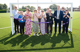A group of 12 dignitaries, men and women, including Witham Mayor Cllr Paul Heath. They are standing in a football goal which had a purple ribbon tied from post to post. A tall gentleman in a suit has just cut the ribon and everyone else is clapping.