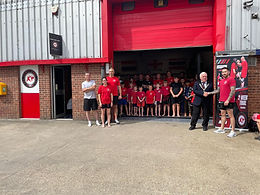 Witham Mayor Cllr Paul Heath, wearing a suit, is shaking hands with a gentleman wearing red tshirt and black shorts. They are standing to the right of a shutter door which is open and a group of children and adults in red tshirts and black shorts are posed in the doorway