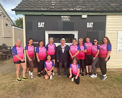 Witham Mayor Cllr Paul Heath stood in fron of a cricket scoreboard. Paul is wearing a suit and his Mayors chains. He is with 11 ladies,2 of whom are kneeling and all are wearing pink t-shirts with purple sleeves, and black shorts or leggings
