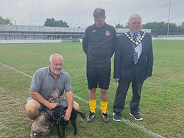 Witham Mayor Cllr Paul Heath wearing a suit and his Mayoral chains. to his left is a gentleman in a Witham Town training kit with yellow socks. Crouched down is a gentleman in Grey t-shirt and beige shorts petting the Mayors two small dogs, one brown one black
