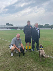 Witham Mayor Cllr Paul Heath wearing a suit and his Mayoral chains. to his left is a gentleman in a Witham Town training kit with yellow socks. Crouched down is a gentleman in Grey t-shirt and beige shorts petting the Mayors two small dogs, one brown one black