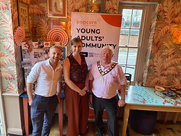 Witham Mayor and Deputy Mayor Cllrs Paul Heath and Jack Coleman stood either side of a lady in a brown top and trousers. They are stood in front of an advertising board which says Popcorn Young Adults Community