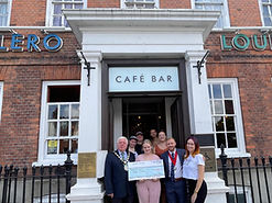 Witham Mayor Cllr Paul Heath, wearing a suit and Mayoral chains, along with the Deputy Mayor also in a suit, with a mixed group of 6 people. The lady in the middle wearing a pink trouser outfit, is holding an oversized cheque