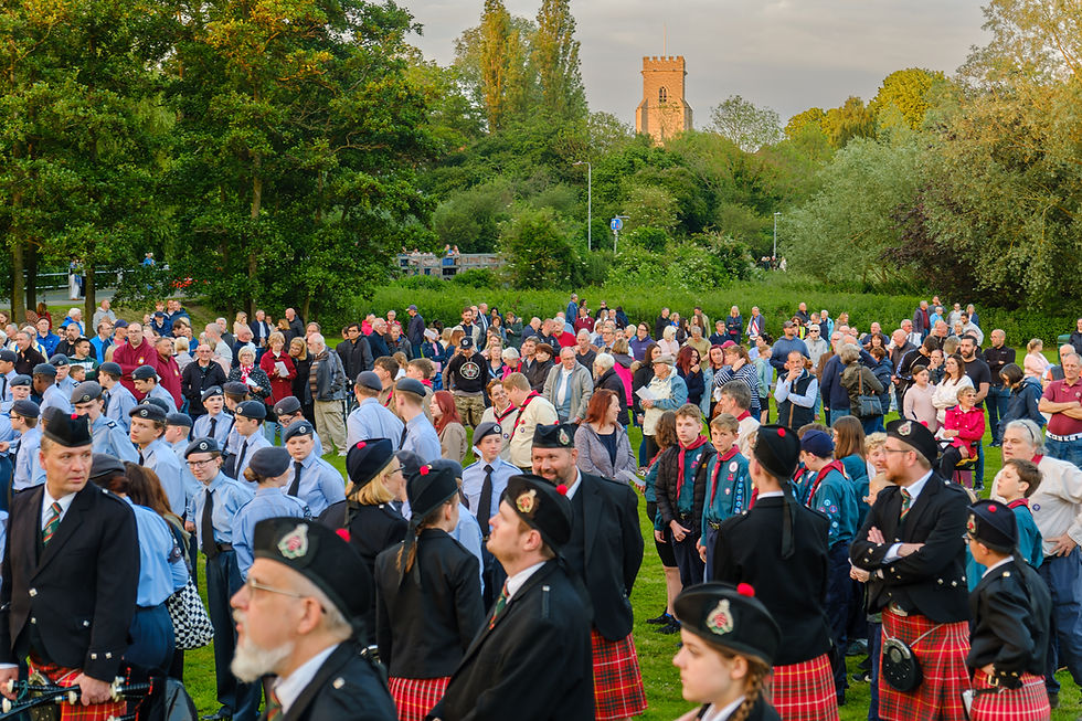 Crowd of people in a park, some in uniforms and kilts, gathering for an event. Trees and St Nicolas Church visible in the background. Mood is lively.