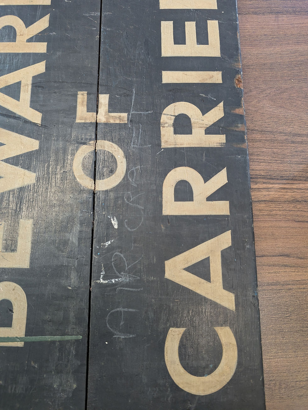 Close up image of black wooden board with beige text "BEWARE OF CARRIER" and faint chalk writing saying Aircraft above it. Background shows a brown wood texture.