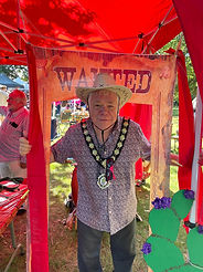 Witham Mayor Cllr Paul Heath under a red gazebo, holding a cardboard frame that says 'Wanted' across the top. Paul is wearing a shirt and trousers, his mayors chains and a cowboy hat