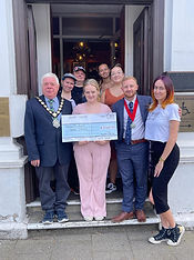 Witham Mayor Cllr Paul Heath, wearing a suit and Mayoral chains, along with the Deputy Mayor also in a suit, with a mixed group of 6 people. The lady in the middle wearing a pink trouser outfit, is holding an oversized cheque