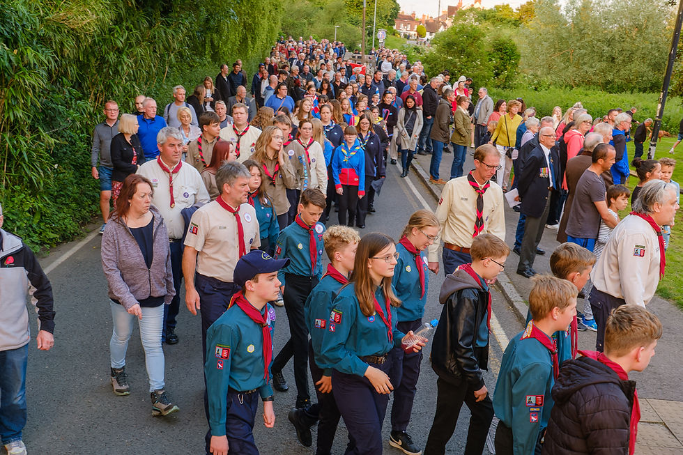 People in scout uniforms and others walk along a road lined with greenery. The mood is organized and focused heading into the River Walk for the D-Day Memorial Ceremony.