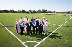 A group of 12 dignitaries, men and women, including Witham Mayor Cllr Paul Heath, stood in a group in the corner of a football pitch. A purple ribbon goes from side to side of the photo which has just been cut while everyone else claps