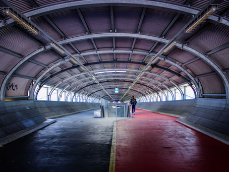 Arched tunnel with red and black paths, lone person walking away. Metal beams above, "P" sign visible. Industrial and isolated mood.