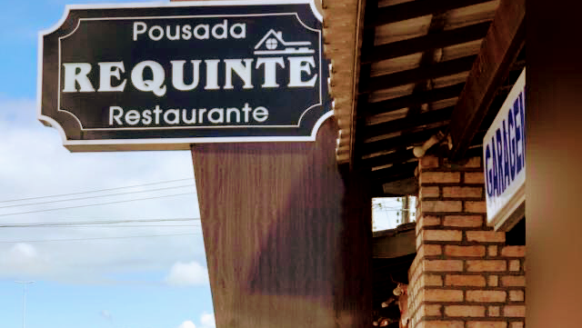 Placa de pousada e restaurante "REQUINTE" em fundo azul. Estrutura de tijolos e madeira ao fundo. Letreiro de garagem à direita. A melhor pousada de Santo Estevão BA