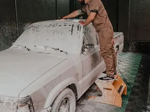Pessoa realizando a esfregação manual de um carro durante a lavagem automotiva, com o veículo completamente coberto por espuma de sabão ativo.
