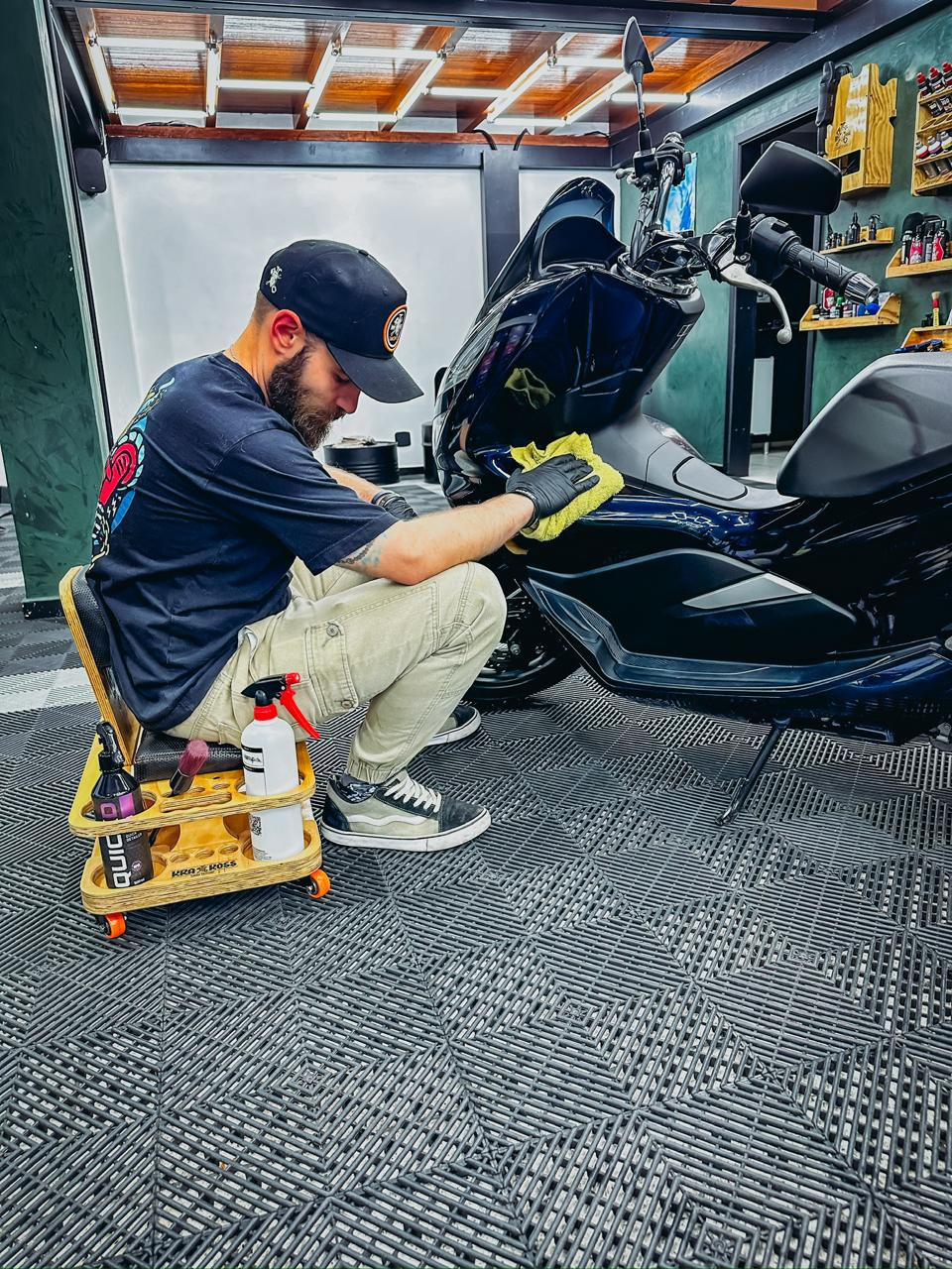 Profissional de detalhamento automotivo utilizando o Banco Detailer 2.0 da Krakoss Garage Solution, sentado ergonomicamente para lavar e detalhar uma moto, otimizando o conforto e a produtividade durante o serviço.