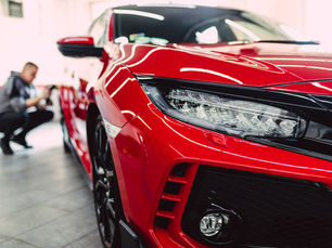 Carro esportivo vermelho em showroom, com close no farol; ao fundo, pessoa agachada fotografa o veículo.
