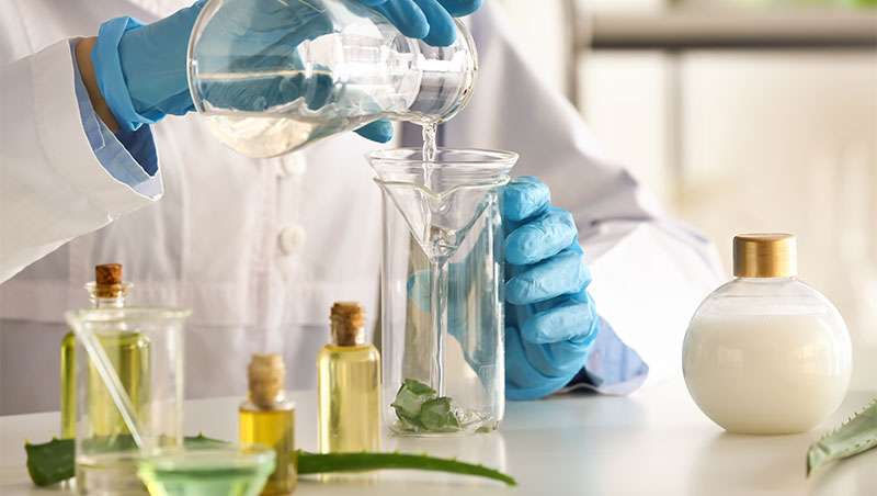 Pessoa de indústria de cosméticos de jaleco e luvas azuis derrama líquido em frasco. Mesas com frascos e folhas. Ambiente claro, atmosfera de ciência e pesquisa.
