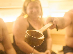 De l’eau versée sur des pierres chaudes crée le löyly dans un authentique sauna finlandais, avec des personnes profitant du sauna en arrière-plan.