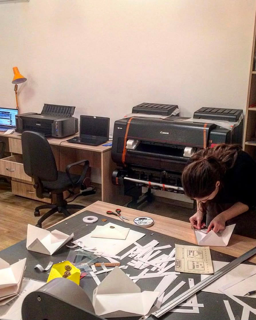 A small print workshop with a large Canon printer, work desks, laptops, paper cutouts, and a person assembling folded paper materials by hand.