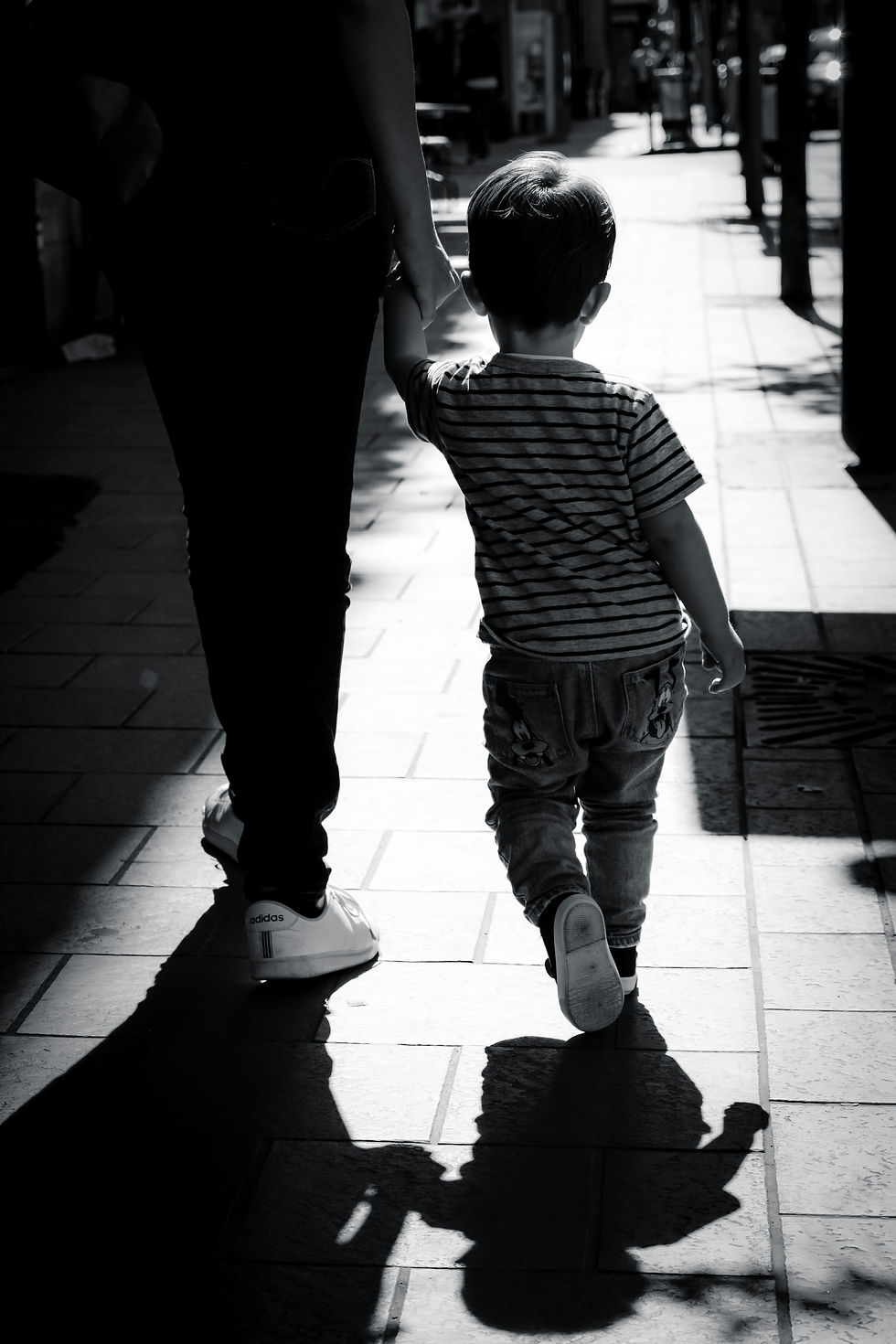 A child and adult holding hands, walking on a sunlit sidewalk. The child wears stripes; shadows are cast. Mood is calm and reflective.