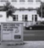 Sign reads "WELCOME TO THE CAPITAL OF SPIES" near street. Car passes by building. Promotes German Spy Museum. Black and white.