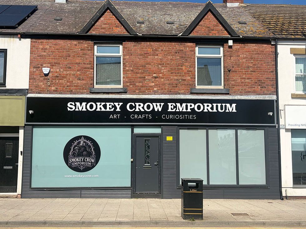 Storefront of Smokey Crow Emporium with a black sign, brick façade, and logo. Text: "Art - Crafts - Curiosities." Sunny street view.