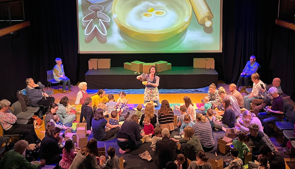 Live storytelling image of Hannah at Cranleigh Arts Centre with family audiences enjoying intreractive play in The Ginger bread Man, with a large screen illustration of baking behind.