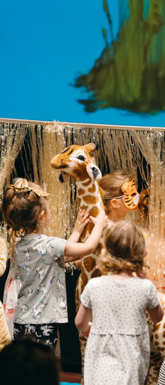 Chldren and Gerald, our large giraffe puppet experience the long wavy grass which is suspended and moved over their heads by the storytelling team.