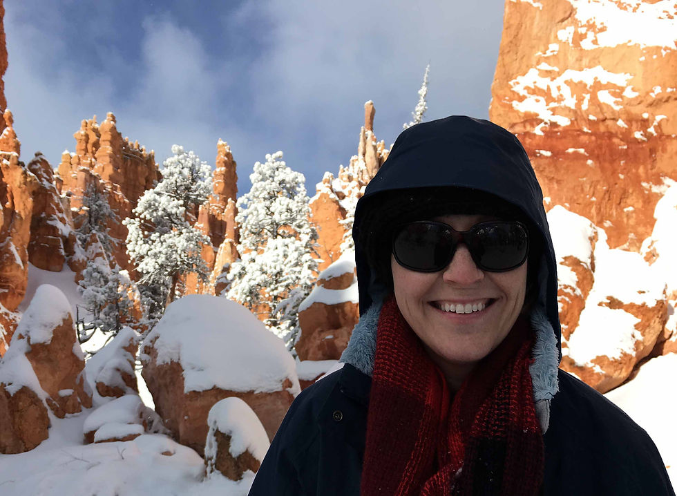 Smiling woman in sunglasses and hooded coat stands amid snow-covered red rock hoodoos under a blue sky near Bryce Canyon National Park Visitor Center.