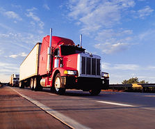 A low-angle shot from the highway surface captures a clean, chrome-accented red semi tractor hauling a white trailer down an open highway on a sunny morning, with a second truck visible approaching from behind. The golden light of early morning glints off the chrome details, with a clear blue sky and soft clouds above a distant tree line. Representing the DOT-regulated, safety-sensitive transportation industry and the federally compliant SAP evaluation services available through Availia TeleHelp PLLC in Spokane and Washington State.