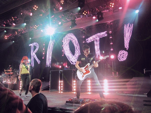 Rock band onstage performing under pink lights, guitarist and singer in front of a large RIOT! backdrop.