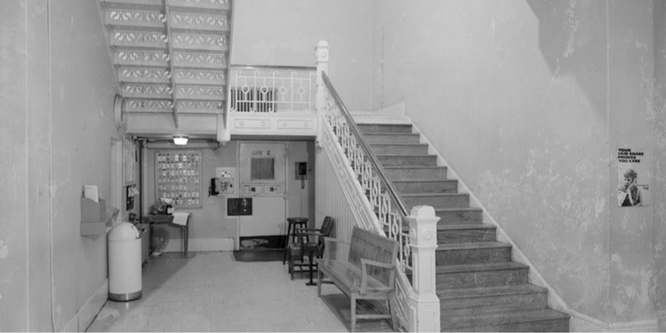 Grayscale stairwell lobby with ornate railings, bench, Gate 2 door, stools, and a wall poster in a quiet old building.