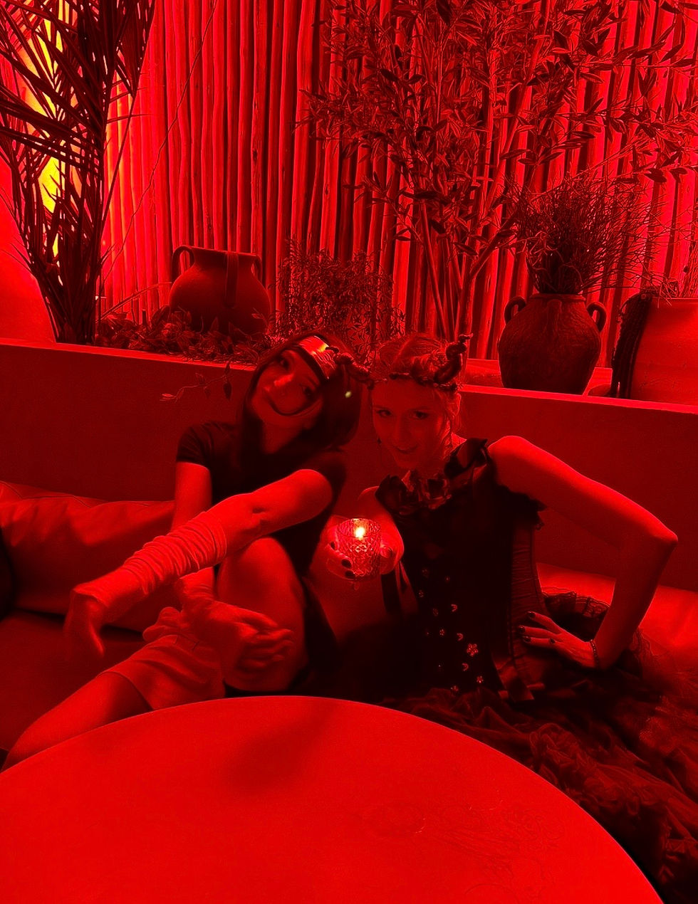 Two women in a red-lit lounge pose on a couch, holding a glowing candle, with tall plants and vases behind them.