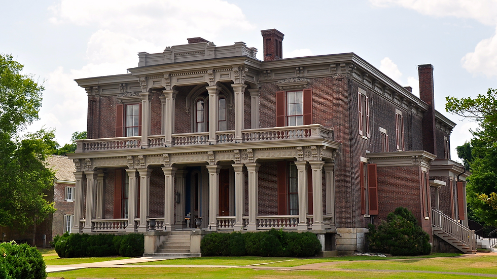 Historic brick mansion with ornate columns, red shutters, and wraparound porches on a manicured lawn under cloudy skies.