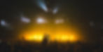 Crowd with raised hands at a concert, bathed in yellow stage lights; screens read ALL I AM IS YOURS.