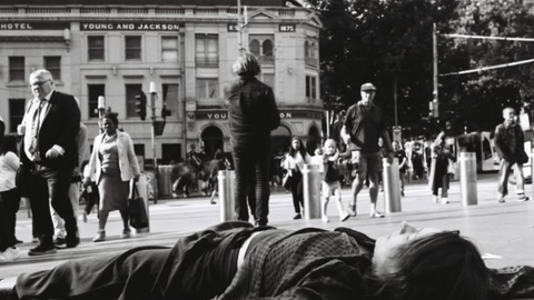 Black and white street photograph by Scott Ransley showing homeless person lying on pavement at Flinders Street Station Melbourne while commuters pass by