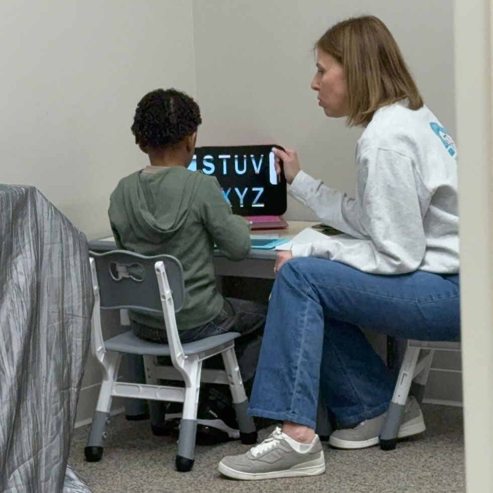 Child and adult at a small table in a classroom. The adult points at a laptop screen displaying letters. Neutral colors, focused mood.