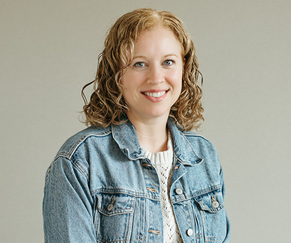 Smiling person with curly hair in denim jacket and white sweater against a plain background.