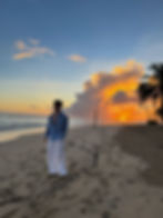 Stanley Dru stood on beach at Zoetry Agua with the sunrise behind him, he is wearing blue baggy shirt and baggy white linen trousers