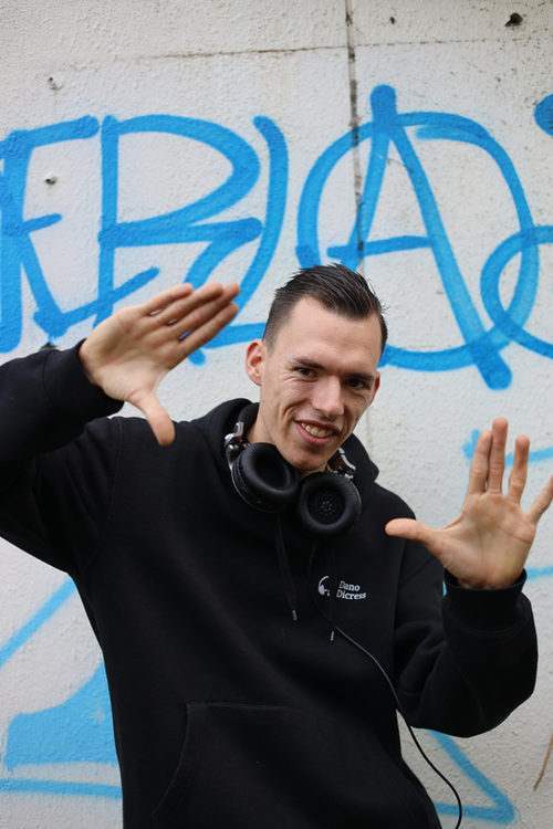 Dano met een koptelefoon om zijn nek voor een muur met blauwe graffiti