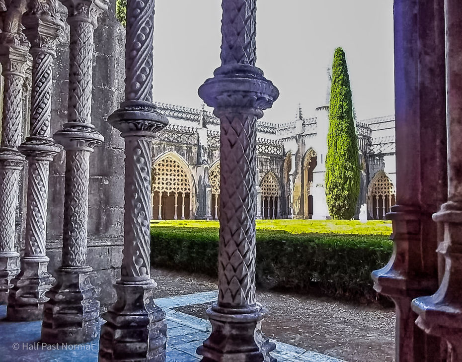 Ornately carved stone columns in the shaded cloisters of Batalha Monastery framing a bright sunlit courtyard with green hedges and Gothic stonework beyond.