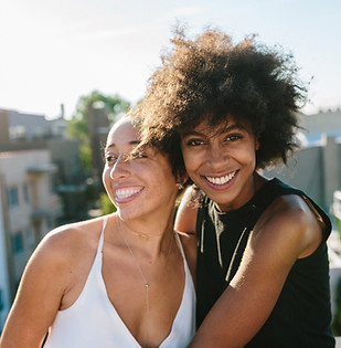 To smiling women of colour embrace while looking comfortably at the camera. This represents YEG Family Counselling's committment to anti-oppression and creating a welcoming and inclusive place for people of colour, neurodivergent people, and LGBTQIA folks.