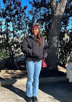 Photo of Erynn Delane, Pen Pal Mentor, standing outdoors among trees wearing a dark puffer jacket, jeans, and a patterned head wrap