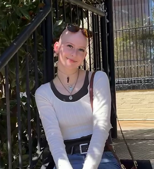 Photo of Melissa McClintock, Pen Pal Mentor, standing outdoors near a fence, wearing a white long-sleeve top and a black vest