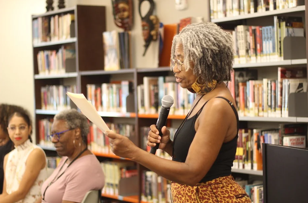 Older Black woman with grey locs holding a microphone and paper to introduce panel