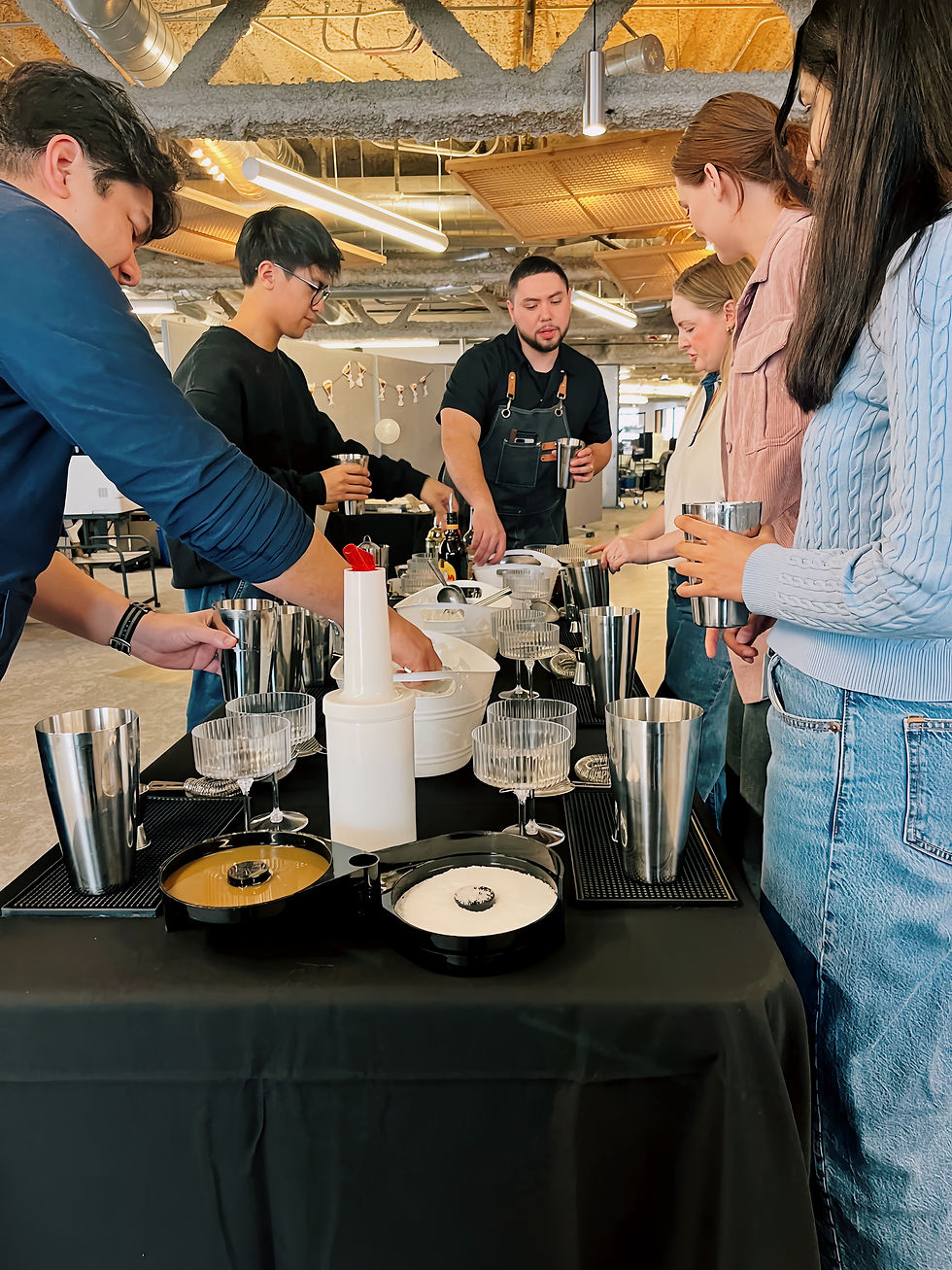 People gather around a table setting with cocktail tools in a modern kitchen. They're engaged, learning mixology with various silver utensils.